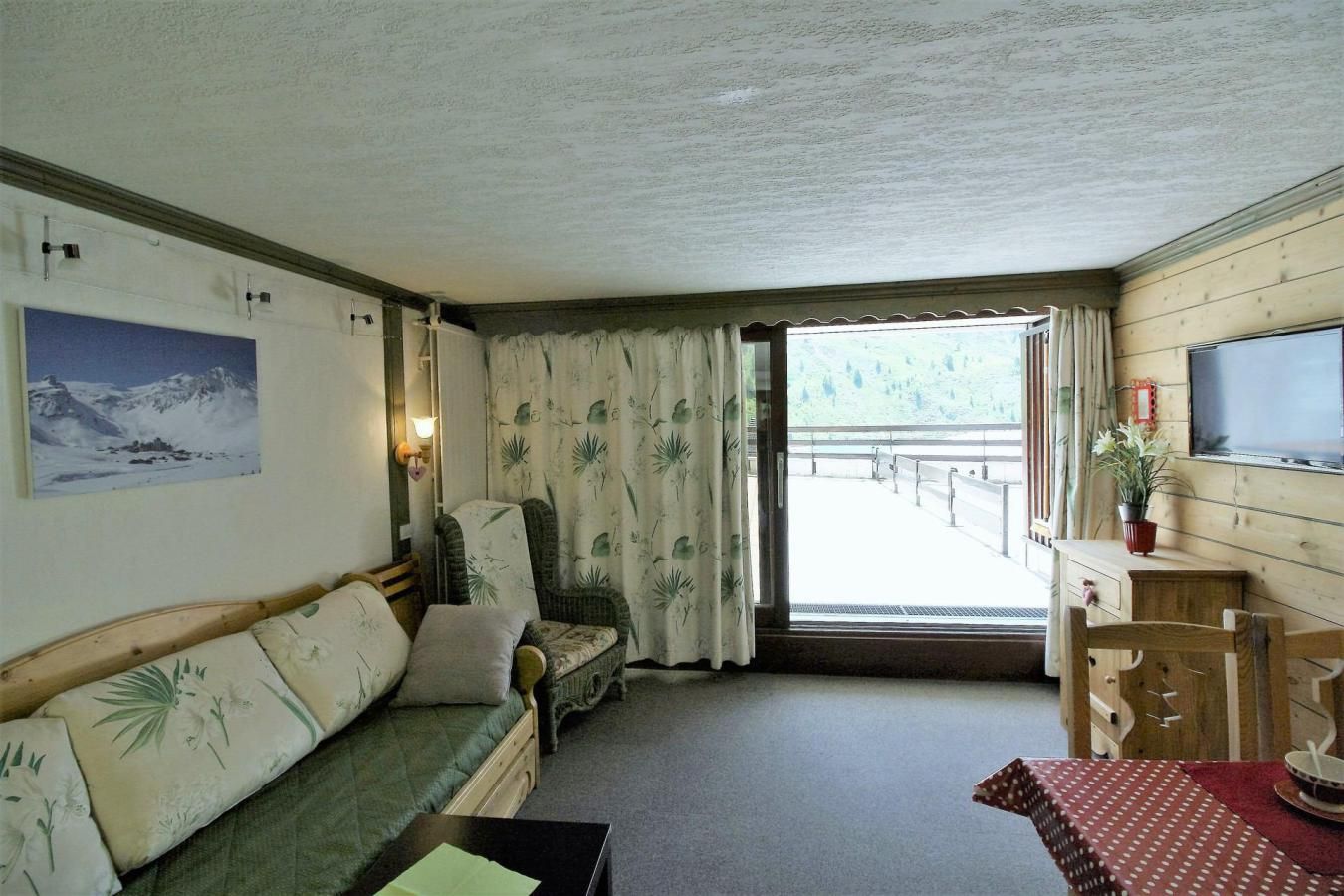 A cozy room with a sofa, a chair, a table, and a TV. There's a window with curtains and a view of a balcony.