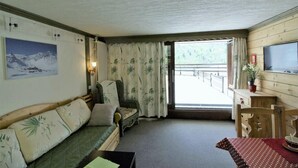 Una habitación acogedora con sofá, sillón, mesa y televisor. Se aprecia una ventana con cortinas y vistas a un balcón.