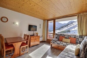A room with a wooden ceiling, a dining table with chairs, a sofa with cushions, a television, and a window with a mountain view. - 10m from ski slopes, 6th floor, balcony, 40m², Méribel-Mottaret (Méribel - Les Allues)
