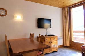 Una mesa de madera con cuatro sillas, un televisor montado en la pared y un ventanal con cortinas.