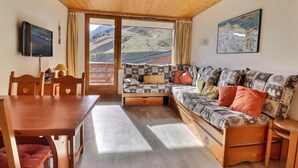 Ein gemütliches Zimmer mit Holzboden, einer Couch mit gemusterten Kissen, einem Fernseher und einem Esstisch mit Stühlen.