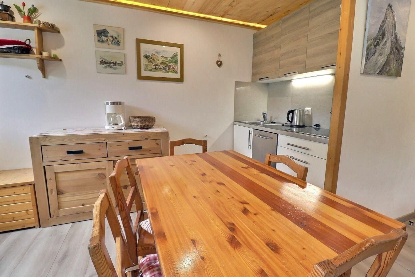 A compact kitchen with a wooden table and chairs, a small cabinet, and a shelf with kitchen items.