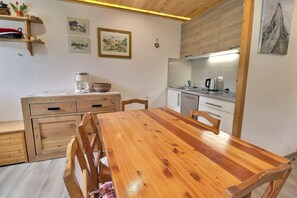 A compact kitchen with a wooden table and chairs, a small cabinet, and a shelf with kitchen items.