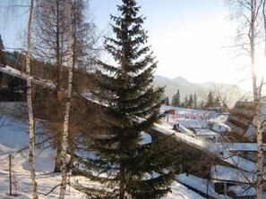 A snow-covered landscape with a tall evergreen tree in the foreground, a mountain range in the background, and a small building with a red roof.