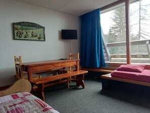 A room with a wooden bench, a bed with a pink pillow, a television on the wall, and a window with blue curtains.