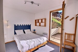 A bedroom with a wooden bed, a bedside table with a lamp, a radiator, and a view of a balcony with a chair.