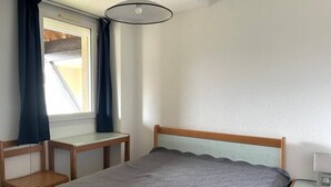 A single-bed room with a wooden headboard, a bedside table, a small wooden chair, and a window with curtains.
