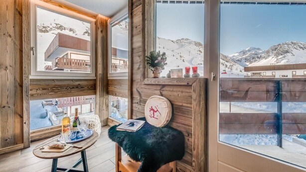 Ein gemütliches Zimmer mit Holzwänden, einem großen Fenster mit Blick auf einen verschneiten Berg, einem kleinen Tisch mit Flasche und Gläsern sowie einem flauschigen Hocker.