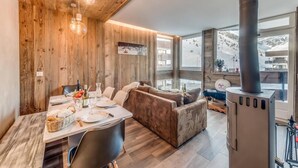 Ein gemütliches Zimmer mit Holzbalkendecke, einer Couch, einem für zwei Personen gedeckten Esstisch und einem großen Fenster mit Blick auf eine verschneite Landschaft.