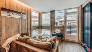 Ein gemütliches Zimmer mit Holzvertäfelung, einem Ledersofa, einem kleinen runden Tisch und Blick auf verschneite Berge durch große Fenster.