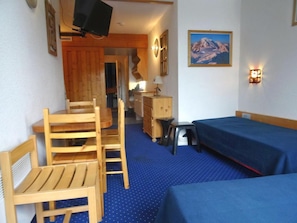 A room with two single beds, a wooden desk with a chair, a small table with a lamp, and a dresser.
