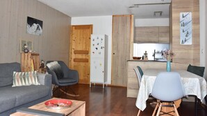 A cozy living room with a grey sofa, a dining table with chairs, a wooden door, and a kitchen area with wooden cabinets and a fridge.