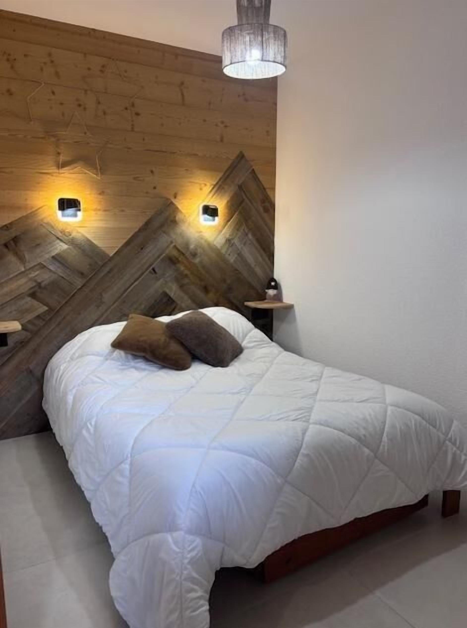 A bedroom with a wooden headboard, two wall-mounted lights, a bed with white bedding, and a bedside table.