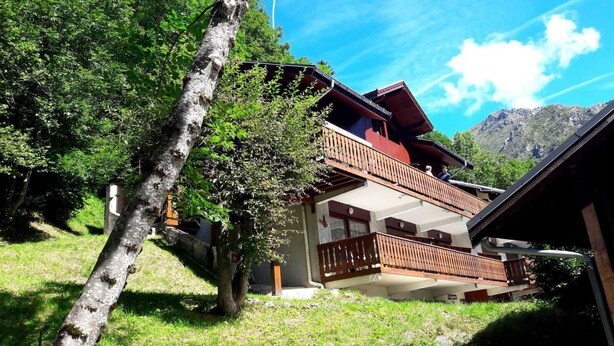 Un chalet à deux étages doté de balcons en bois, entouré d’une végétation luxuriante et avec des montagnes en toile de fond.