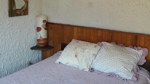 Une chambre avec un plafond en bois, un lit recouvert d’une literie à motifs floraux, une table de chevet avec un vase, et un mur orné d’un tableau encadré.