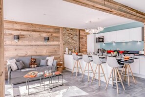 Una cocina moderna con armarios blancos, un comedor con sillas grises y un sofá gris con un peluche de oso.