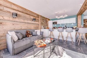 Una sala moderna con un sofá gris, un muro decorativo de madera, una zona de cocina con armarios blancos y una barra con taburetes.