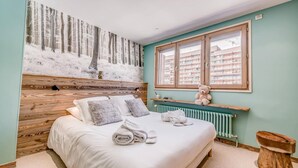 Une chambre avec une tête de lit en bois, une grande fenêtre donnant sur des immeubles, un ours en peluche sur une étagère et des serviettes soigneusement pliées sur le lit.