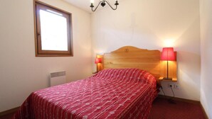 A bedroom with a wooden headboard, a red patterned bedspread, two bedside lamps, a window, and a wall-mounted heater.