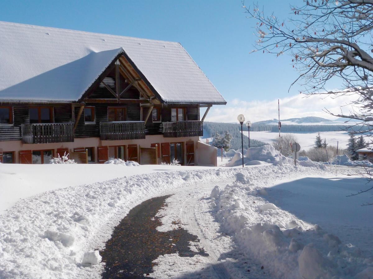 A lodge with a snow-covered roof and a balcony. A snow-covered path leads to the entrance.