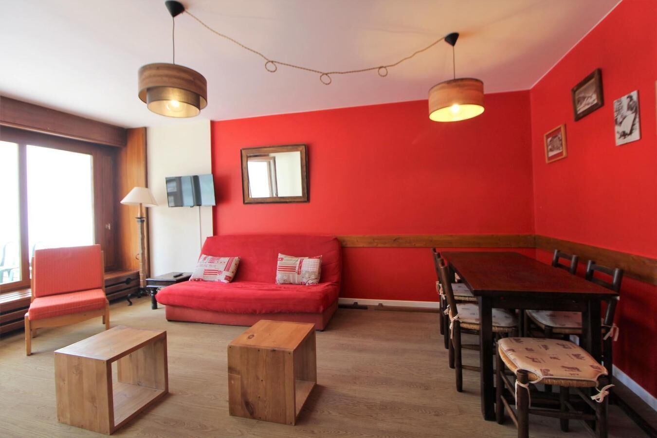Une pièce avec un mur rouge, un canapé en bois, une table à manger avec des chaises et deux petites tables d’appoint en bois.