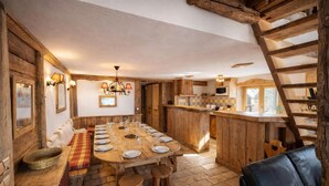 Une cuisine rustique avec une grande table à manger en bois, dressée pour un repas, des poutres en bois au plafond et un sol en pierre.