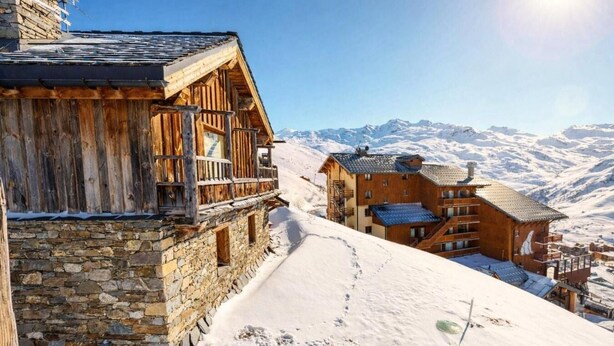 Un chalet en bois aux murs de pierre, doté d’un balcon et offrant une vue sur des montagnes enneigées.