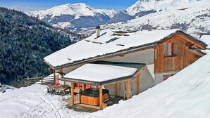 Un chalet de madera con techo nevado, una terraza de madera y un jacuzzi en primer plano.