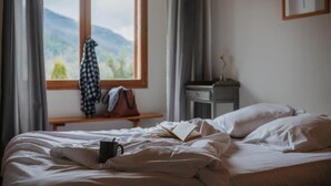 Um quarto com cama, janela com vista para montanhas, mesa de cabeceira com abajur e um quadro na parede.