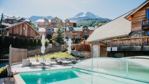 Área de piscina com espreguiçadeiras e guarda-sóis, cercada por edifícios de madeira com varandas, com montanhas ao fundo.