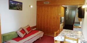 A room with a wooden wall, a bed with red and white bedding, a table with a newspaper-covered tablecloth, and a TV mounted on the wall.