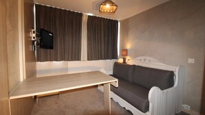 Une petite pièce chaleureuse, avec une table en bois, un canapé gris, un téléviseur fixé au mur et une lampe de chevet.
