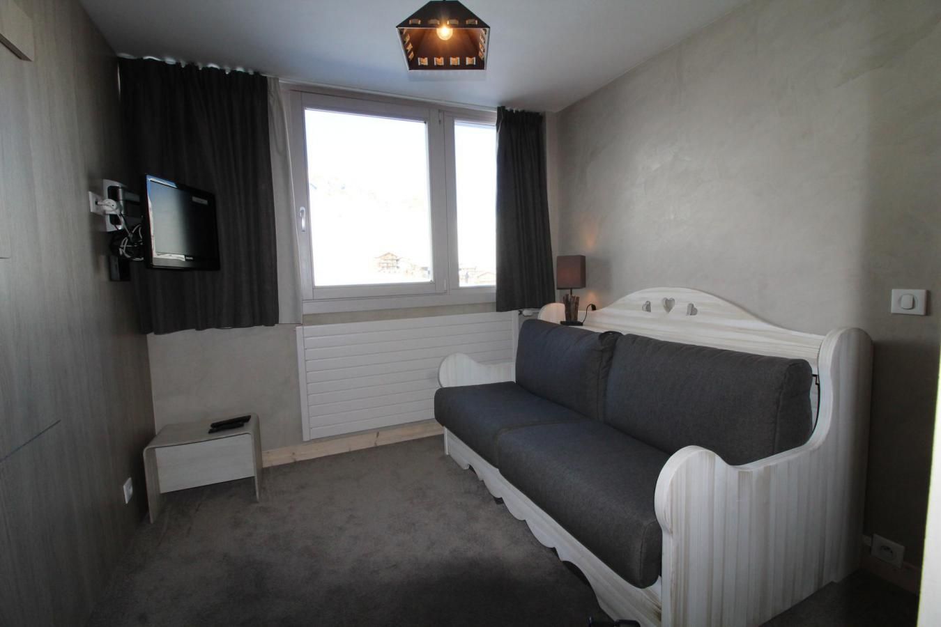A small, modern room with a white sofa, a flat-screen TV mounted on the wall, a small bedside table, and a window with curtains.