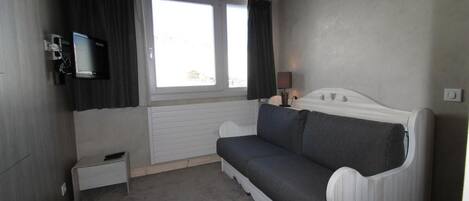 A small, modern room with a white sofa, a flat-screen TV mounted on the wall, a small bedside table, and a window with curtains.