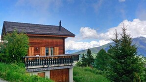 A wooden chalet with a balcony, surrounded by lush greenery and mountains in the background.