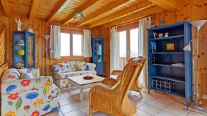 A cozy wooden cabin interior with a sofa set, a dining table, wicker chairs, and a blue bookshelf.