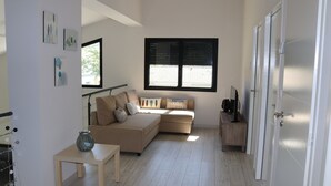 En moderne stue med en beige hjørnesofa, et lille træbord og en glasvase. Der er et stort vindue med persienner, en dør og en trappe, der fører til et andet niveau.