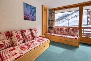 A cozy room with a wooden sofa and bench, a large window with a view of a snowy mountain landscape, and a wall-mounted picture of an anemone. - 10m from ski slopes, 1st floor, balcony, 27m², Méribel-Mottaret (Méribel - Les Allues)