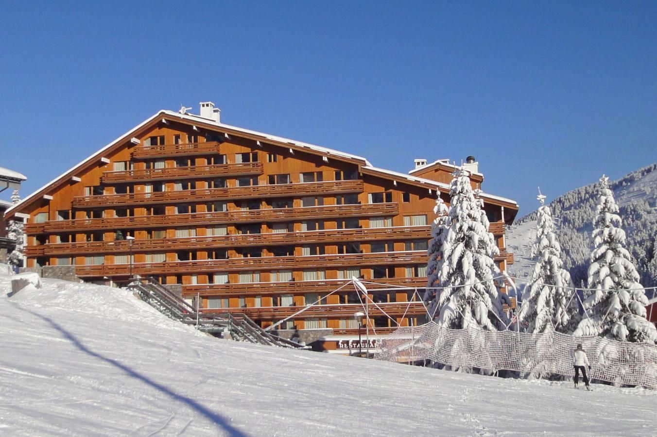 A multi-story wooden building with balconies, likely a hotel, surrounded by snow-covered trees and a ski slope.