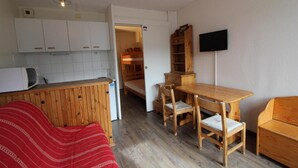 Una habitación compacta con kitchenette, una zona de comedor con muebles de madera, un televisor montado en la pared y un sofá a rayas rojas.
