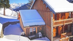 A wooden chalet with a snow-covered roof, a balcony, and a view of snow-covered mountains.