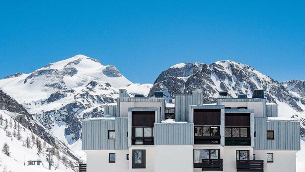 Een modern gebouw met een symmetrische gevel, grote ramen en een plat dak, op de achtergrond besneeuwde bergen en een strakblauwe lucht.