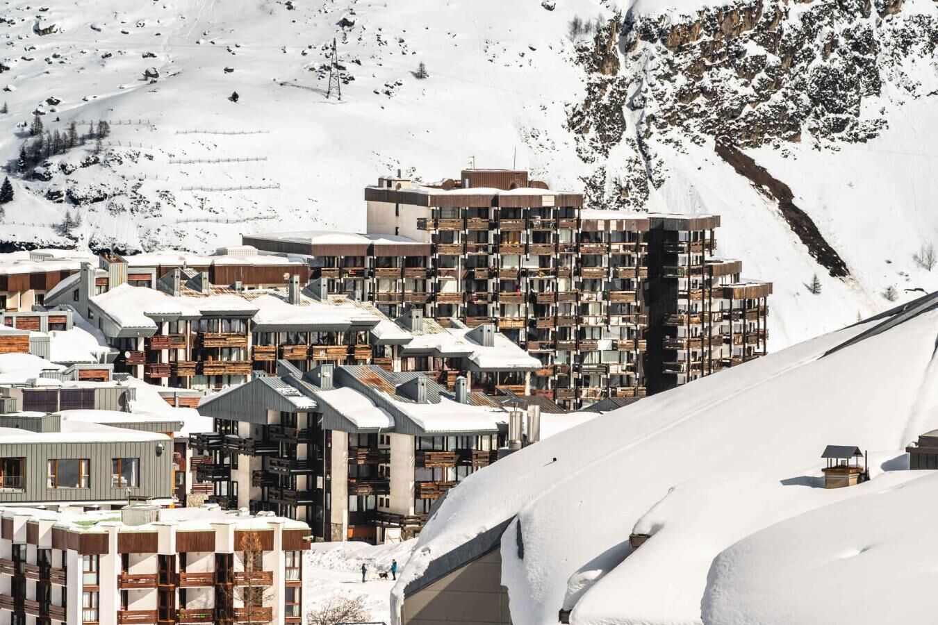 A snow-covered mountain town with modern multi-story buildings, some with balconies, and a ski slope in the background.
