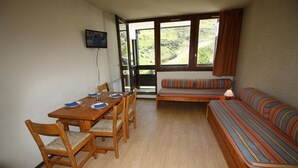 Een kamer met een houten eettafel voor vier personen, een bank met gestreepte kussens en een televisie aan de muur.