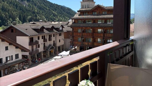A balcony with a table set for two, overlooking a town with traditional buildings and a prominent church tower. - 3rd floor, balcony, tv, 26m², Le Grand Bornand (Le Grand-Bornand)