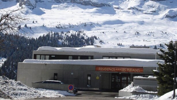 A building labeled "Residence Les Pleiades" with a parking area and snow-covered mountains in the background.