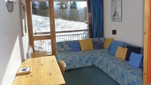 Een gezellige kamer met een houten tafel, een geruit bankje en een groot raam met uitzicht op een besneeuwd landschap.