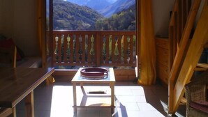 A room with a view of mountains through a large window, a wooden table, and a wicker chair. - 4th floor, swimming pool, balcony, tv, ski locker, 37m², Valloire (Valloire)