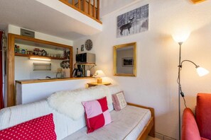 Ein gemütliches Wohnzimmer mit einer Couch, einer Lampe und einem roten Sessel. Im Hintergrund ist eine Küchenzeile mit einer Mikrowelle und einer Kaffeemaschine erkennbar.