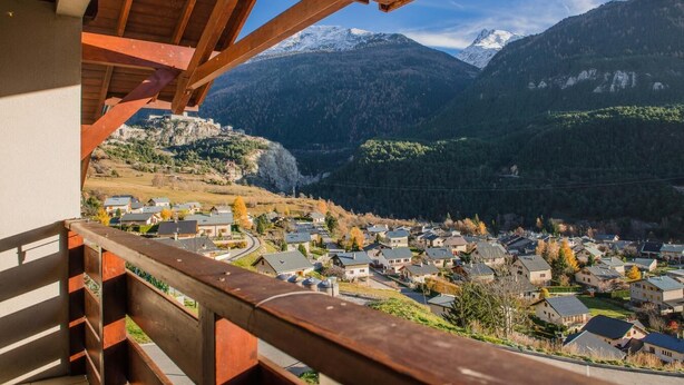A balcony with a wooden railing overlooking a village and snow-capped mountains. - Parking, tv, ski locker, 35m², La Norma (Avrieux)